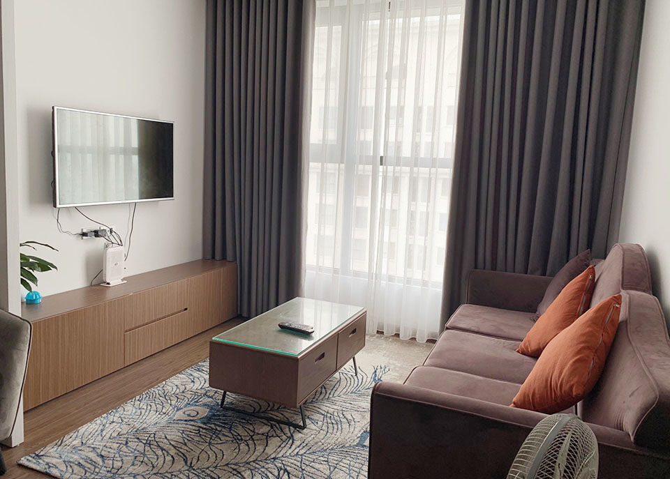 Phòng khách căn hộ chung cư với chiếc tivi treo tường, bàn ghế sofa, gối ôm màu cam, thảm trải sàn màu trắng xanh