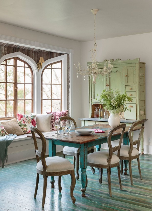 hình ảnh phòng ăn phong cách đồng quê với bàn gỗ màu xanh dương pastel, tủ lưu trữ xanh lá nhạt, cửa sổ kính dạng vòm, đèn chùm và bình hoa trang trí, ghế ngồi bên cửa sổ, gối tựa hoa văn màu sắc