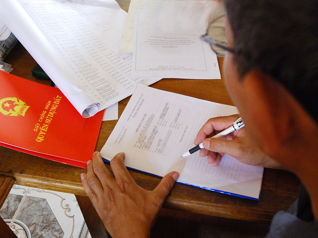 một người đàn ông đang đọc bản hợp đồng, bên cạnh là sổ đỏ đất